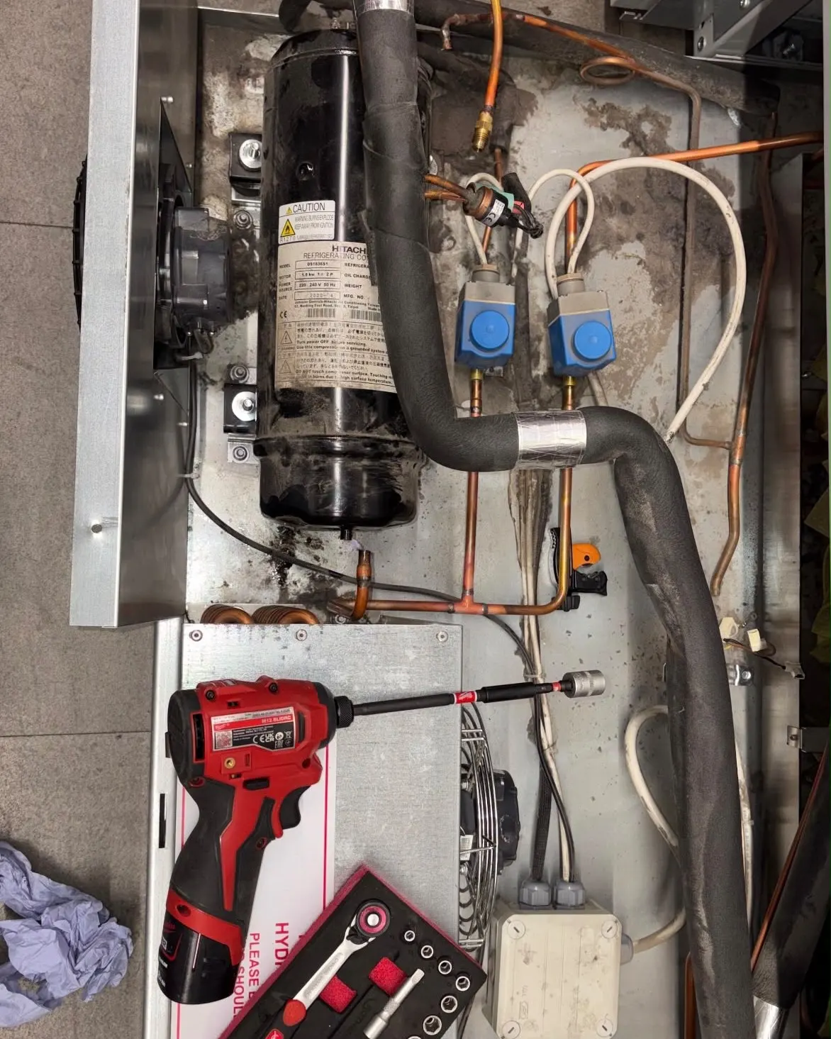 Compressor and pipework inside a commercial freezer cabinet during a compressor repair, with a drill and socket set