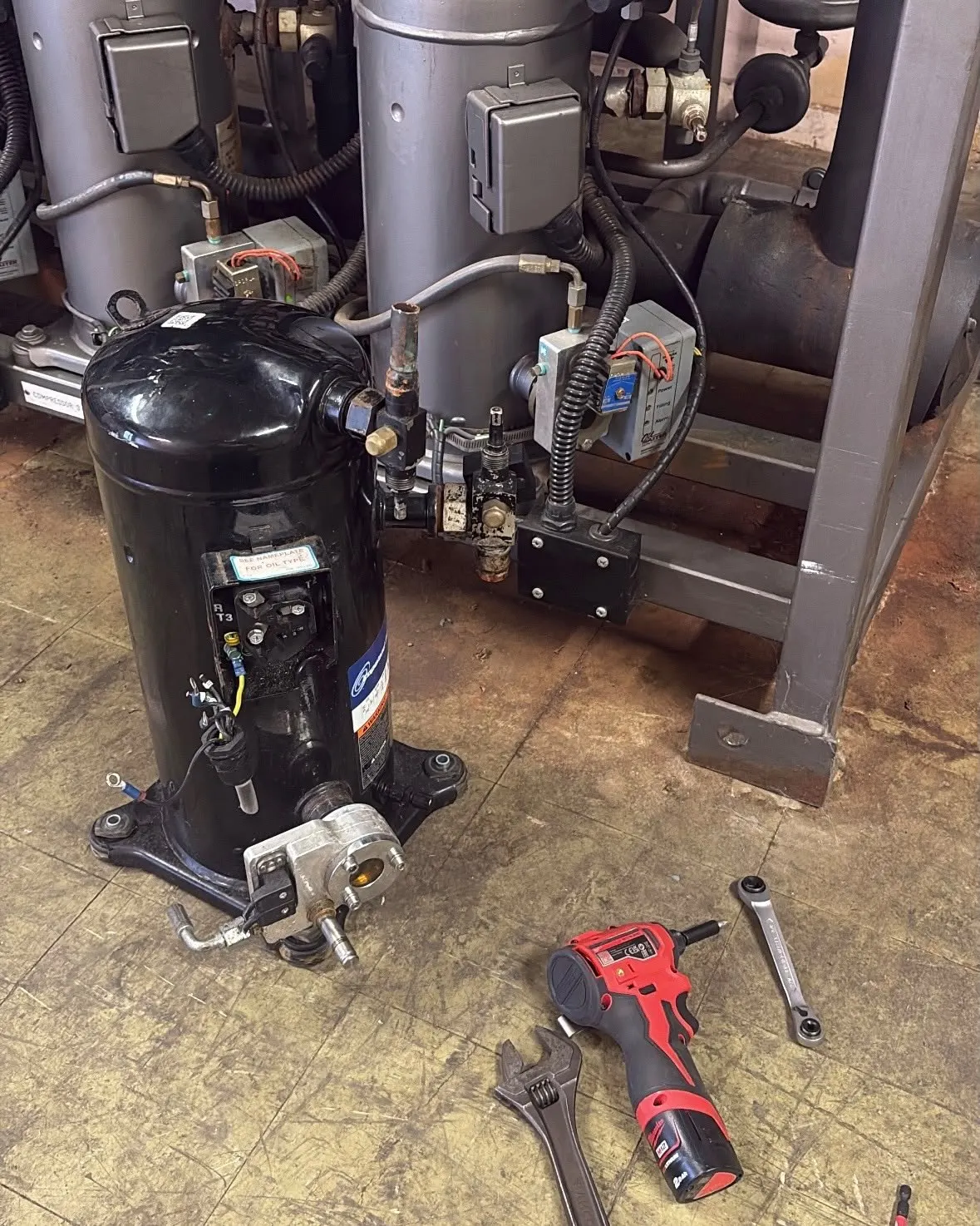 New scroll compressor on the floor beside a refrigeration compressor pack, with tools ready