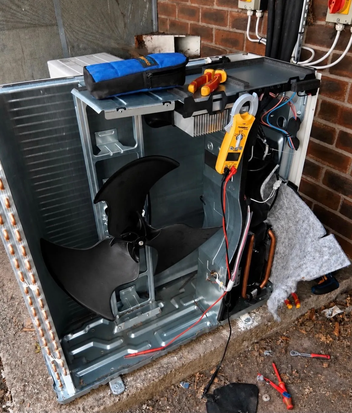 Outdoor air conditioning unit with its casing removed for repair, with a clamp meter and hand tools on the unit
